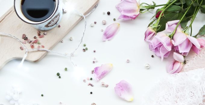 white ceramic mug filled with black liquid on beige chopping board beside pink roses on white surface